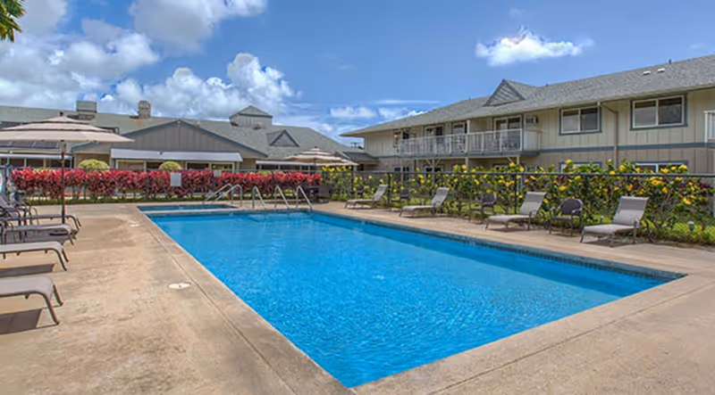 Outdoor swimming pool with lounge chairs around it, surrounded by a fence and flowering bushes. In the background, there are two-story buildings under a partly cloudy blue sky.