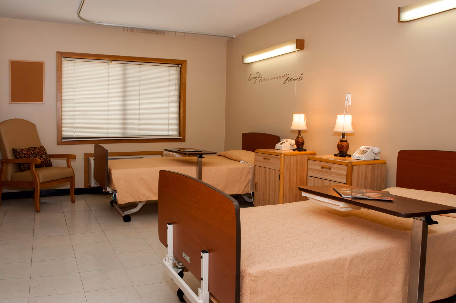 A room with two hospital-style beds covered with beige bedspreads, each with an overbed table. There are two wooden nightstands between the beds, each with a lamp and a telephone. A cushioned armchair with a patterned pillow is placed near a window with closed blinds. The walls are beige, and there is a motivational quote on the wall above the nightstands. The floor is tiled.