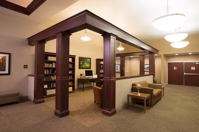 A warm communal lounge with wooden columns, seating, bookshelves, a computer desk, and pendant lighting.