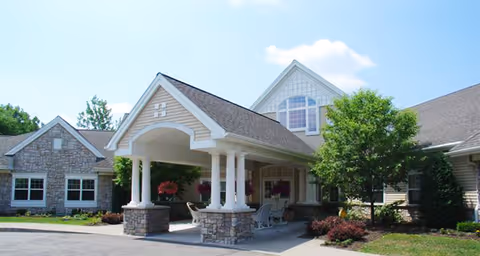 Exterior view of a senior living facility with a covered entrance supported by white columns and stone bases. The building has beige siding, stone accents, and multiple windows. There is a small tree and landscaped bushes near the entrance under a partly cloudy sky.