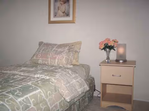 A small bedroom with a patterned bed, nightstand, flowers, and framed wall art.