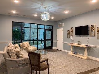 A cozy living room area with a beige sofa, two cushioned armchairs, a wooden coffee table, and a patterned rug. The room has a large glass door and windows leading outside, a wall-mounted flat-screen TV, and decorative wall art. The walls are painted light blue with white trim, and a modern chandelier hangs from the ceiling.
