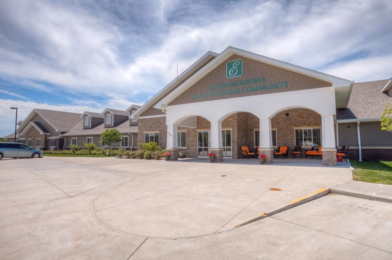 Exterior view of Edencrest at Green Meadows assisted living community building with a covered entrance, outdoor seating with orange cushions, and a large concrete driveway under a partly cloudy sky.