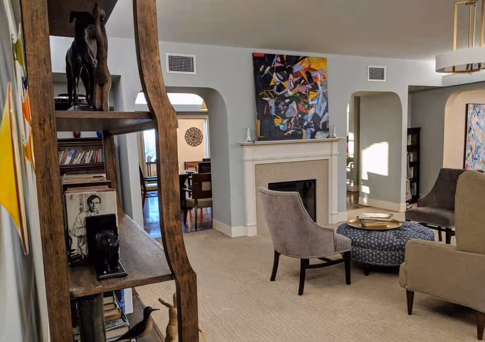 A cozy living room area featuring a fireplace with a colorful abstract painting above it. The room has a beige carpet, a round blue patterned ottoman, and several upholstered chairs arranged around it. To the left, there is a wooden bookshelf with various books and decorative items. The walls are painted light gray, and there are arched doorways leading to other rooms.