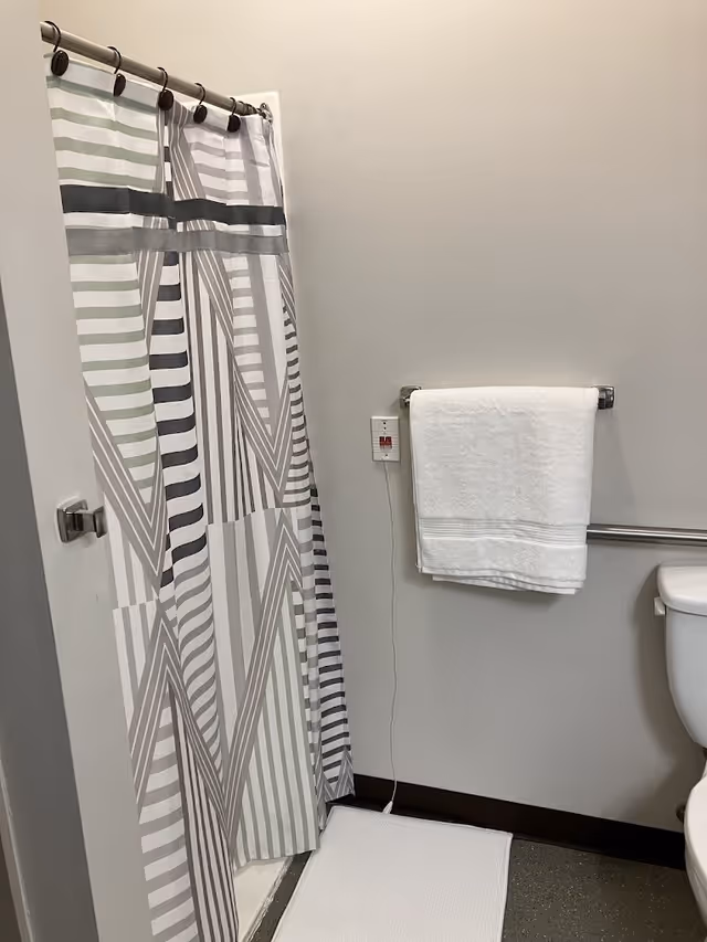 A bathroom corner featuring a shower with a striped shower curtain in shades of gray and green. A white towel hangs on a metal towel rack mounted on a light gray wall. Below the towel rack is a white bath mat on a dark floor. A toilet is partially visible on the right side. There is also an emergency pull cord with a red button on the wall next to the towel rack.