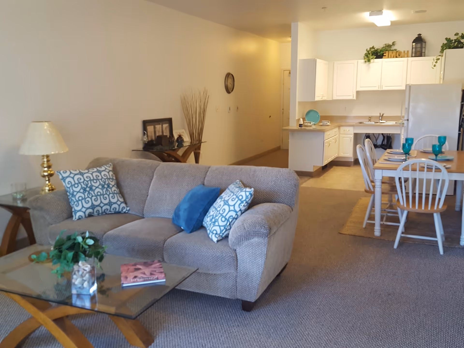 A cozy living area with a beige sofa adorned with blue and patterned pillows, a glass coffee table with a plant and a book, a side table with a lamp, and a dining table set with turquoise glasses and plates. The background shows a white kitchen with cabinets, a sink, and a refrigerator.