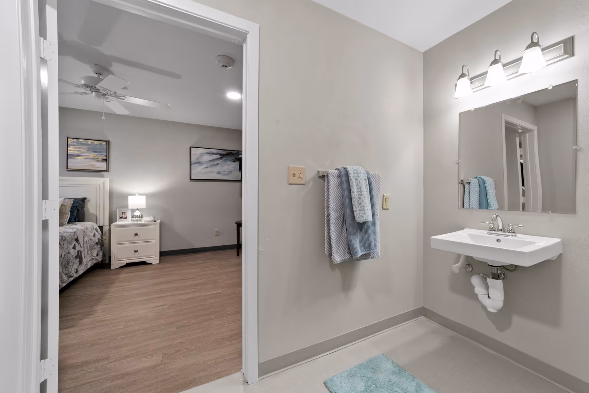 View of a bathroom with a wall-mounted sink, mirror, and three-light fixture above. Towels hang on a rack next to the sink, and a blue bath mat is on the floor. Through an open door, a bedroom is visible with a bed, nightstand, lamp, and wall art.