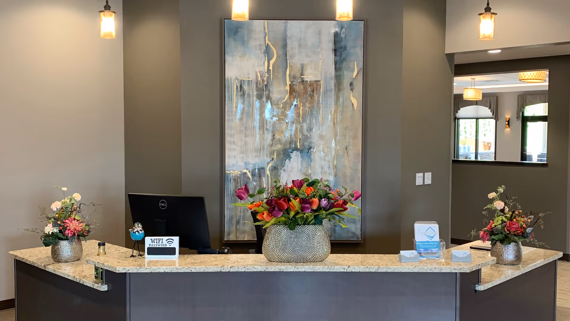 Reception desk area with a granite countertop featuring three flower arrangements in decorative vases. Behind the desk is a computer monitor and a large abstract painting on the wall. The space is well-lit with hanging pendant lights and has a neutral color scheme with gray walls.