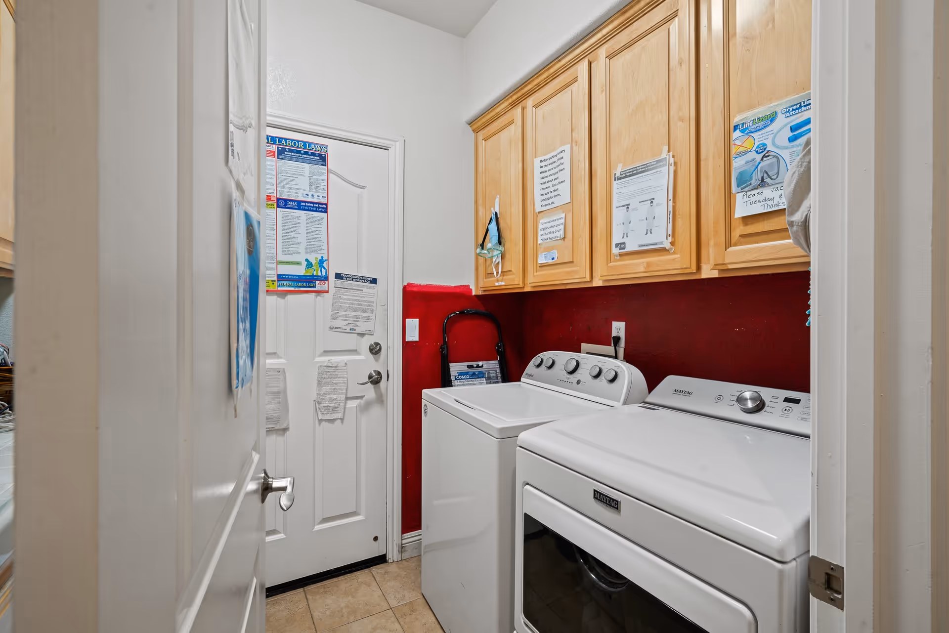 Laundry room with a white washing machine and dryer side by side, light wooden cabinets above them, and various notices and instructions posted on the cabinets and door. The walls are painted white and red, and there is a small step ladder leaning against the wall.