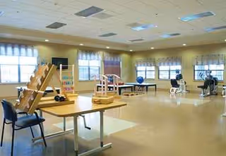 Bright therapy gym with ramps, tables, and residents seated by large windows.
