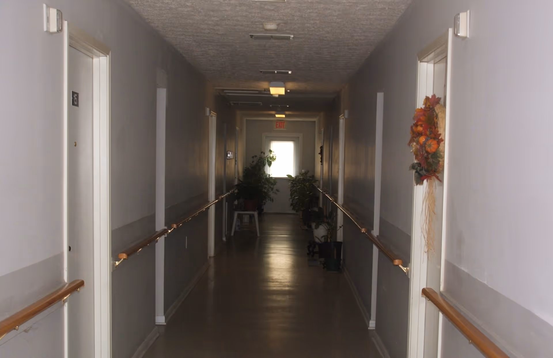 Long interior hallway with handrails, doors on both sides, a decorative wreath, and plants at the far end.