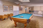 Interior view of a recreational room featuring a pool table with billiard balls arranged on it. The room has carpeted flooring, beige walls adorned with framed artwork, and a seating area with two brown armchairs. A railing separates the pool table area from a hallway in the background.