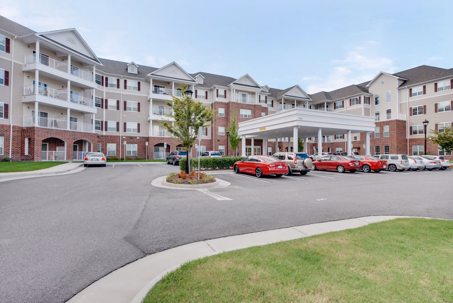 Front exterior of a multi-story senior living building with a covered entrance/porte-cochere and parked cars in the driveway.