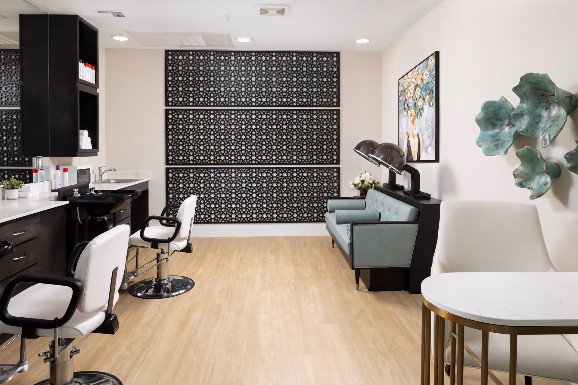 Interior view of a salon area in a senior living facility featuring two white salon chairs with black armrests in front of a black sink and countertop. The room has light wood flooring, a teal cushioned bench with two vintage hair dryers, decorative black patterned panels on the back wall, a floral painting on the right wall, and green abstract wall art. A white chair and a small round table with a gold base are also visible in the foreground.