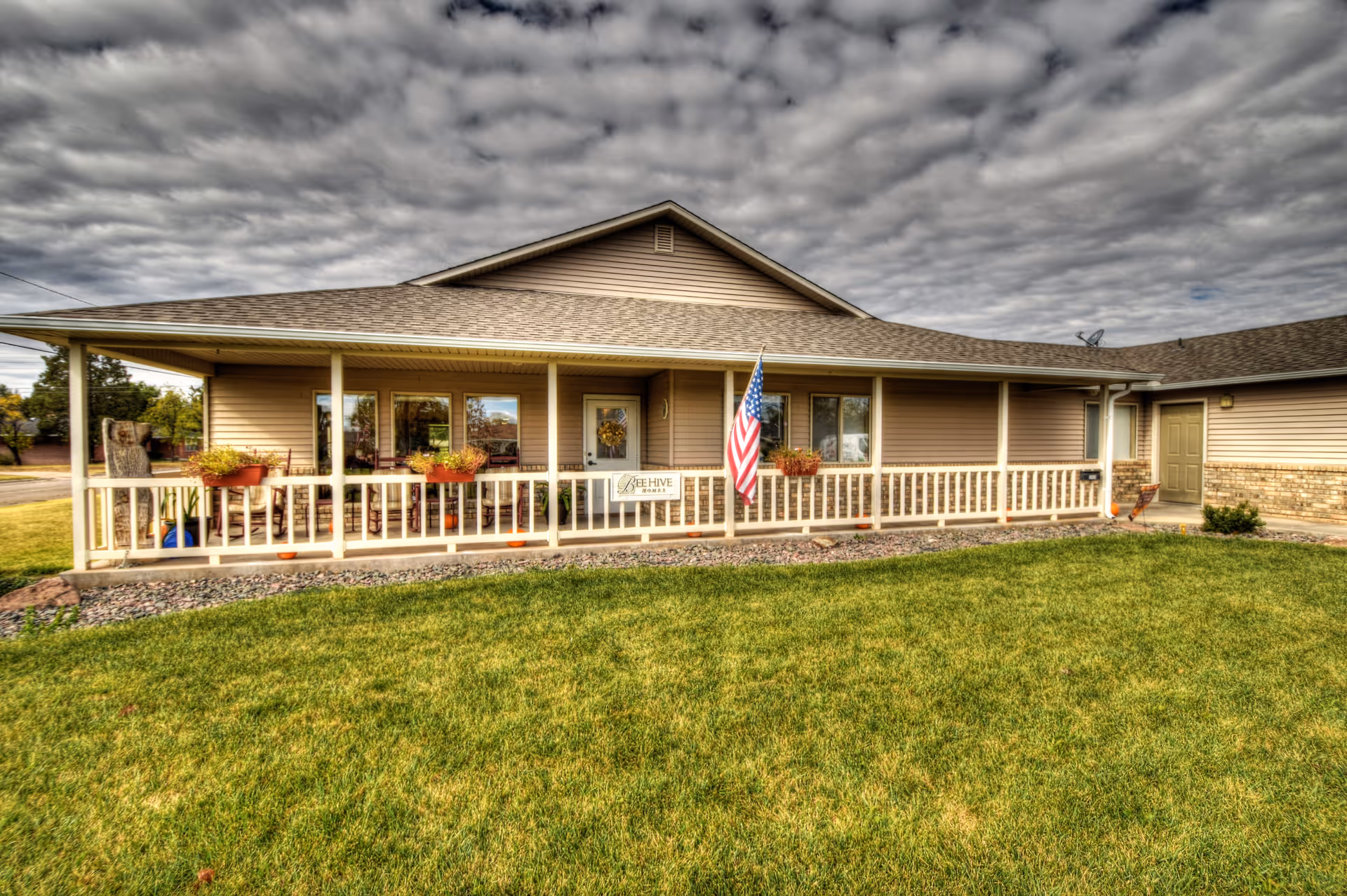 Exterior view of a single-story building with beige siding and a covered porch. The porch has white railings, several potted plants, and an American flag mounted near the entrance door. The building is surrounded by a well-maintained green lawn under a cloudy sky.