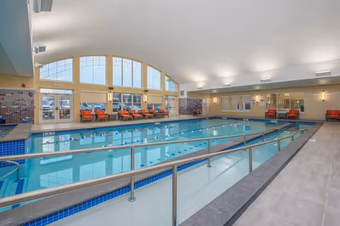 Indoor swimming pool with lane markings, metal handrails, seating along large windows, and a high arched ceiling.