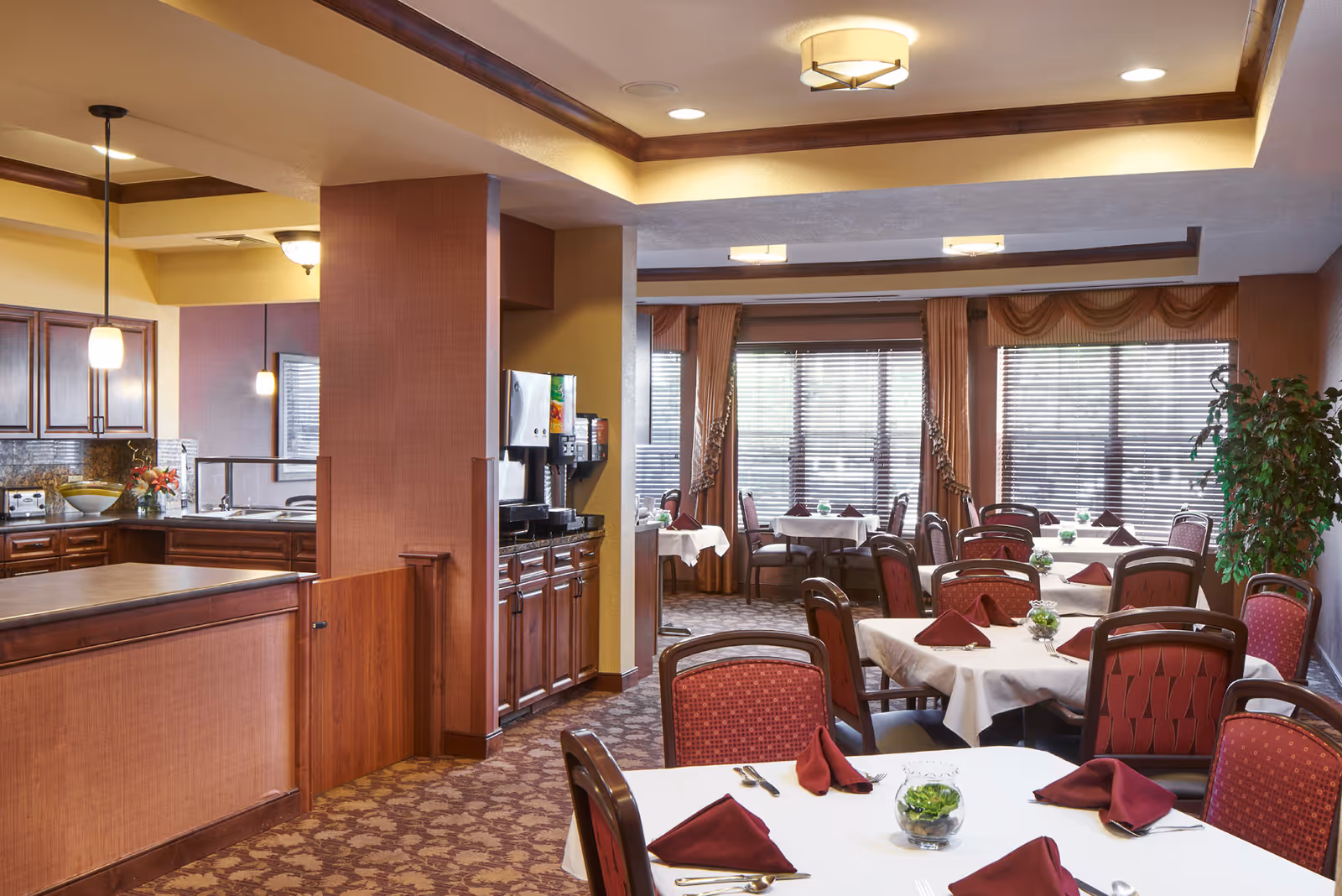 A dining room in a senior care community with multiple tables covered with white tablecloths and burgundy napkins. The room has large windows with blinds and curtains, patterned carpet, and wooden chairs with red upholstery. To the left, there is a kitchen area with wooden cabinets, a countertop, and a beverage dispenser.