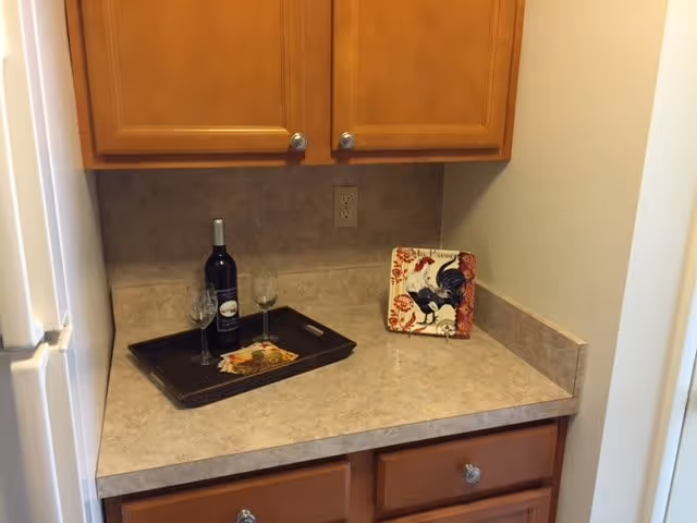 A small kitchen countertop area with light brown cabinets above and below. On the countertop, there is a black tray holding a bottle of wine and two wine glasses. Next to the tray is a decorative plate with a rooster design. The wall behind the countertop has a beige tile backsplash and an electrical outlet.