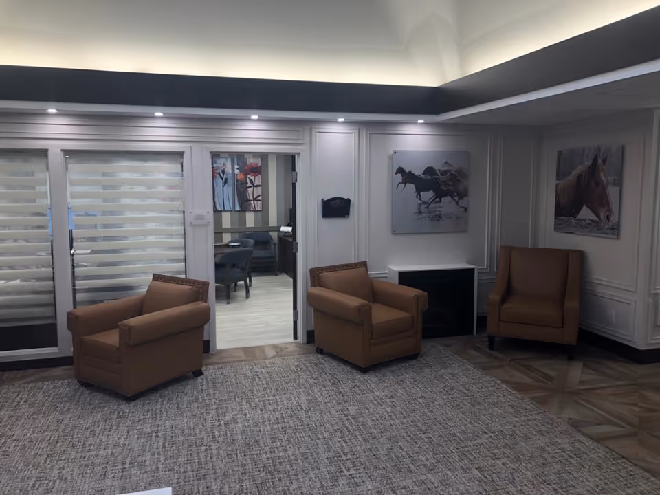 Well-lit common lounge with three tan armchairs, carpeted floor, horse artwork on the walls, and a doorway into a dining area.