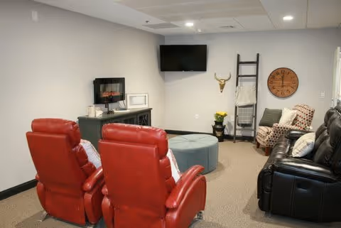 A cozy living room with two red recliners facing a black TV mounted on a light gray wall. There is a black console table with a small electric fireplace and decorative items. A round blue ottoman sits in the center. On the right side, there is a black leather sofa with pillows, a wooden clock on the wall, a decorative bull skull, and a ladder-style blanket rack with a blanket. The room has beige carpet and soft lighting.