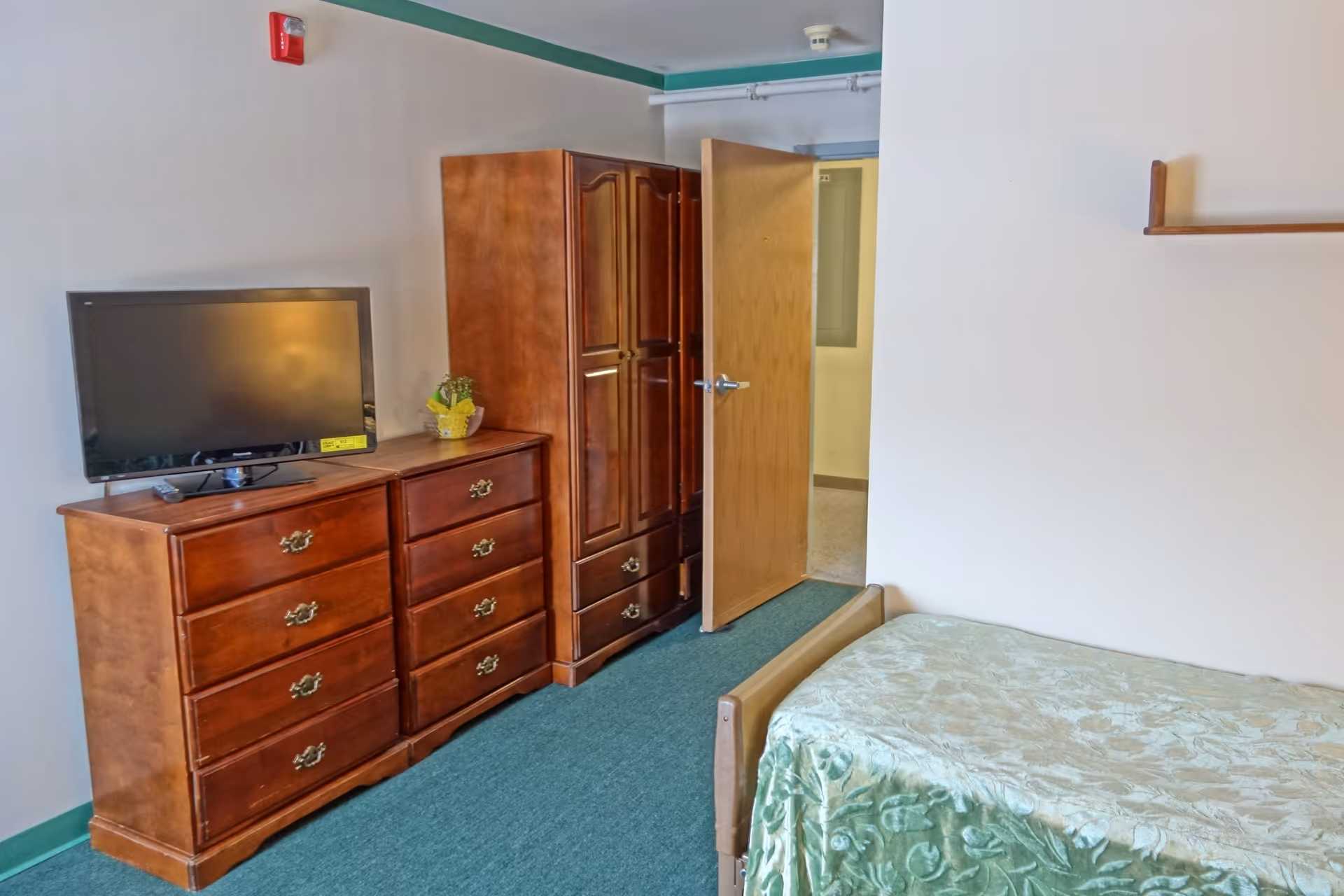 A senior living facility bedroom with a wooden dresser and wardrobe, a flat-screen TV on top of the dresser, a single bed with a green patterned bedspread, an open wooden door, and a small shelf mounted on the wall.