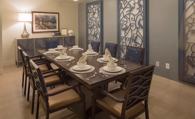 A dining room with a long wooden table set for six people, each place setting includes a plate, bowl, napkin folded in a decorative manner, and silverware. The chairs have dark wooden frames with patterned blue cushions. Behind the table is a sideboard with a lamp, framed artwork of a tree-lined path, and decorative items. The walls have decorative metal panels with intricate designs.