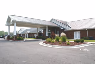 Exterior front entrance of a single-story brick building with a covered porte-cochere, circular driveway, and landscaped beds.