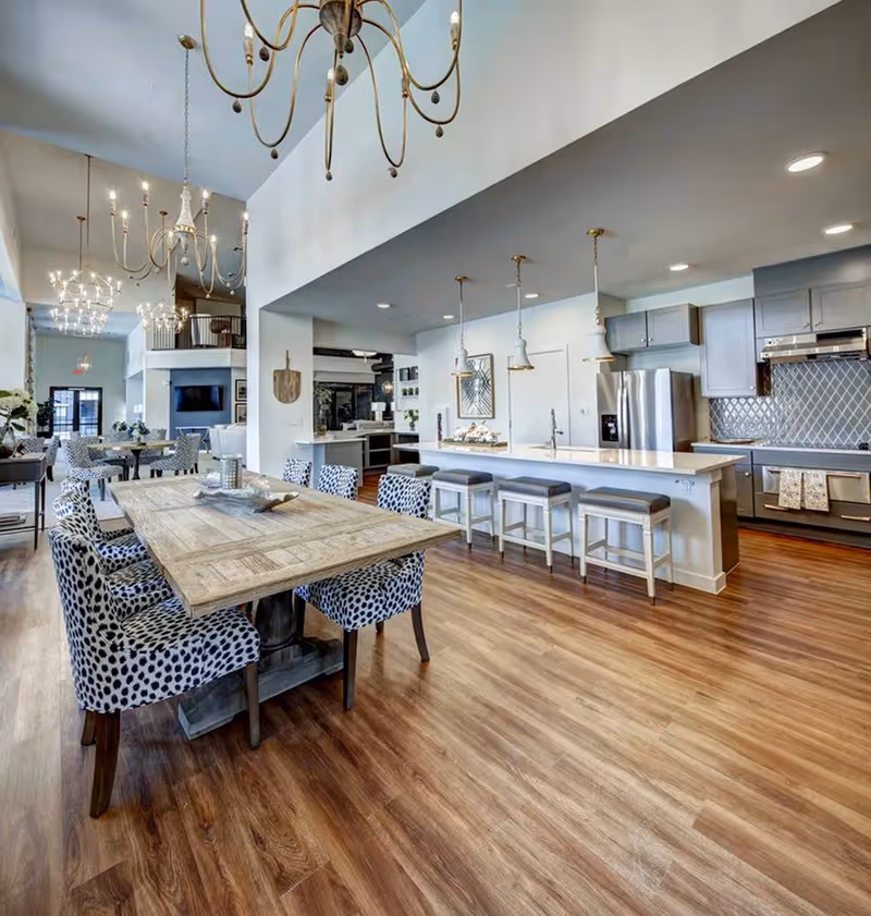 Spacious and modern open-concept dining and kitchen area with wooden flooring. The dining area features a large wooden table surrounded by six upholstered chairs with a black and white spotted pattern. The kitchen has a long white island with four cushioned bar stools, stainless steel appliances, gray cabinetry, and pendant lighting. In the background, there are additional dining tables and chairs, chandeliers, and a fireplace with a mounted TV above it.