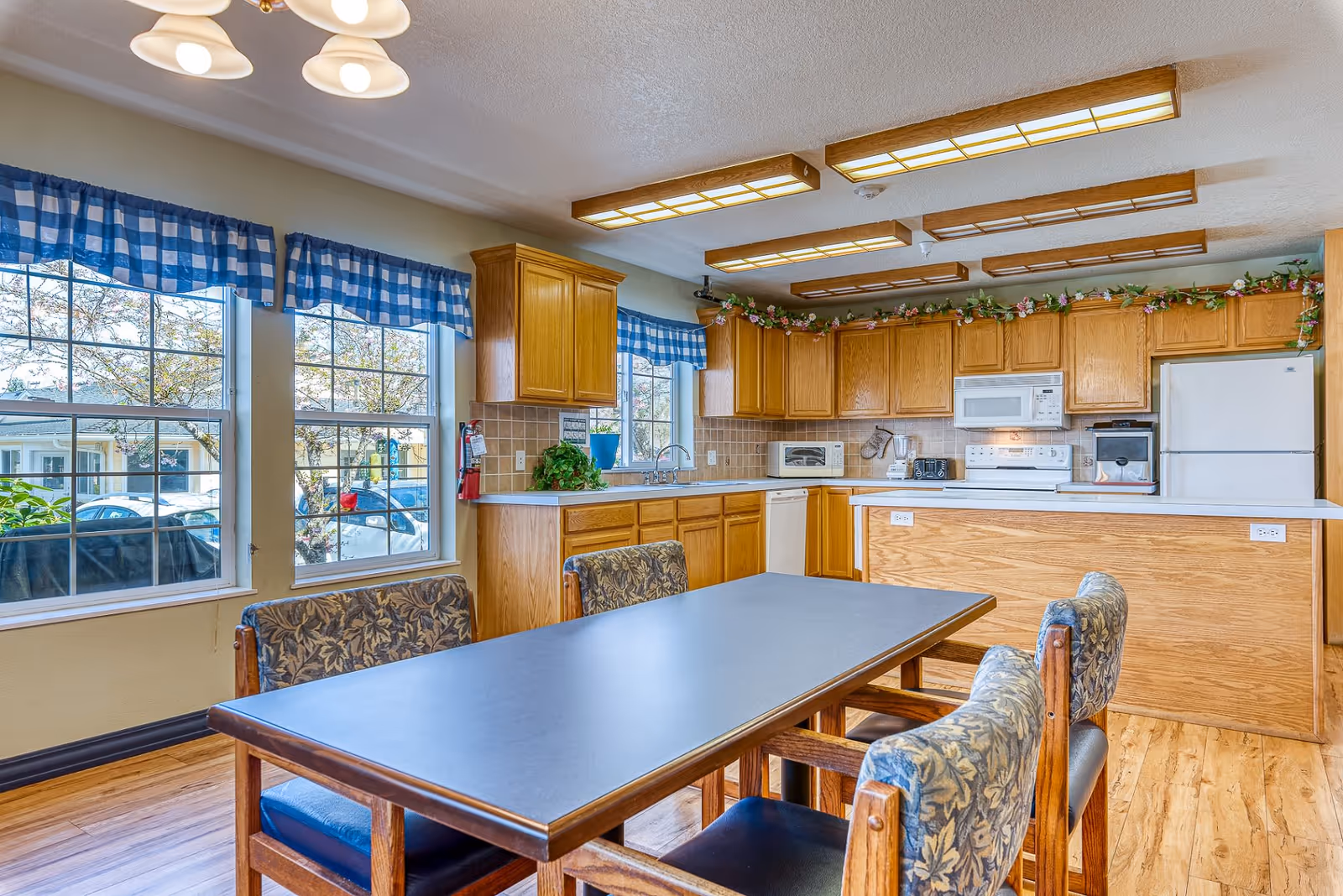 A bright kitchen and dining area with wooden cabinets, a white refrigerator, microwave, stove, and dishwasher. A rectangular dining table with four cushioned chairs is in the foreground. Large windows with blue checkered valances let in natural light, and floral garlands decorate the top of the cabinets.