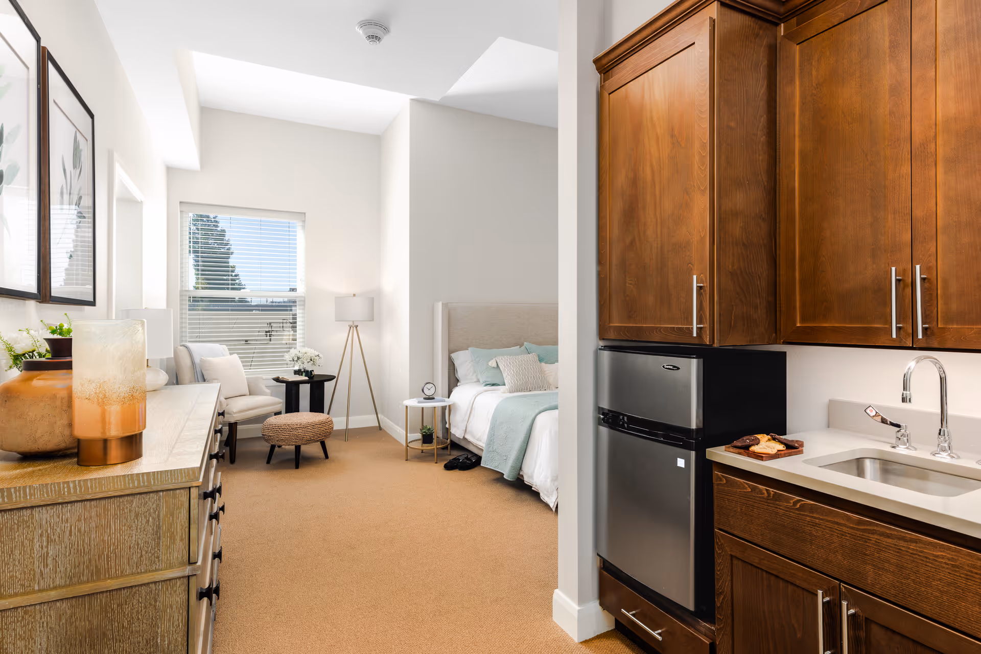 A bright and cozy studio apartment featuring a small kitchenette with wooden cabinets, a stainless steel mini refrigerator, and a sink. The room includes a comfortable bed with light-colored bedding and pillows, a side table with a clock and plant, a floor lamp, a cushioned chair, a small round table, and a woven ottoman. The walls are decorated with framed botanical prints, and a window with blinds allows natural light to fill the space.