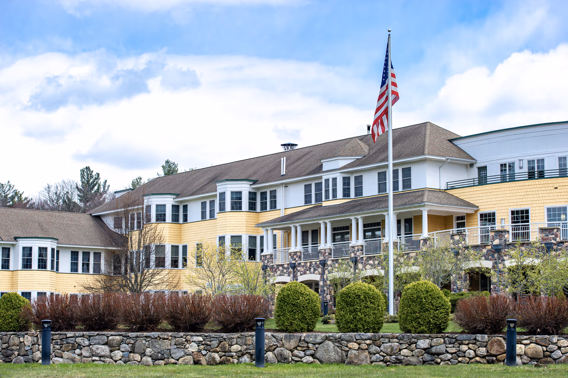Exterior view of a large senior living facility building with yellow siding and white trim, featuring multiple windows and a covered porch. In front of the building is a stone wall, manicured bushes, and an American flag on a flagpole. The sky is partly cloudy.
