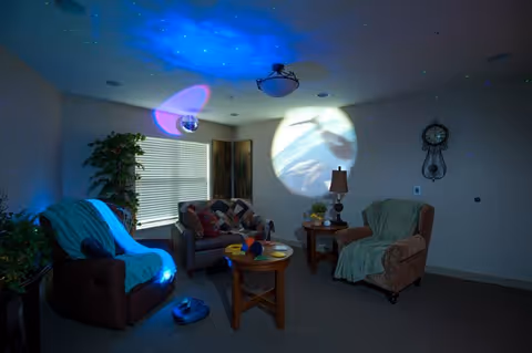 A cozy living room with two armchairs and a sofa, each covered with blankets. A round wooden coffee table is in the center with some items on it. The room is dimly lit with colorful light projections on the ceiling and wall, creating a calming atmosphere. There is a window with blinds, a potted plant, a floor lamp, a wall clock, and a decorative screen in the corner.