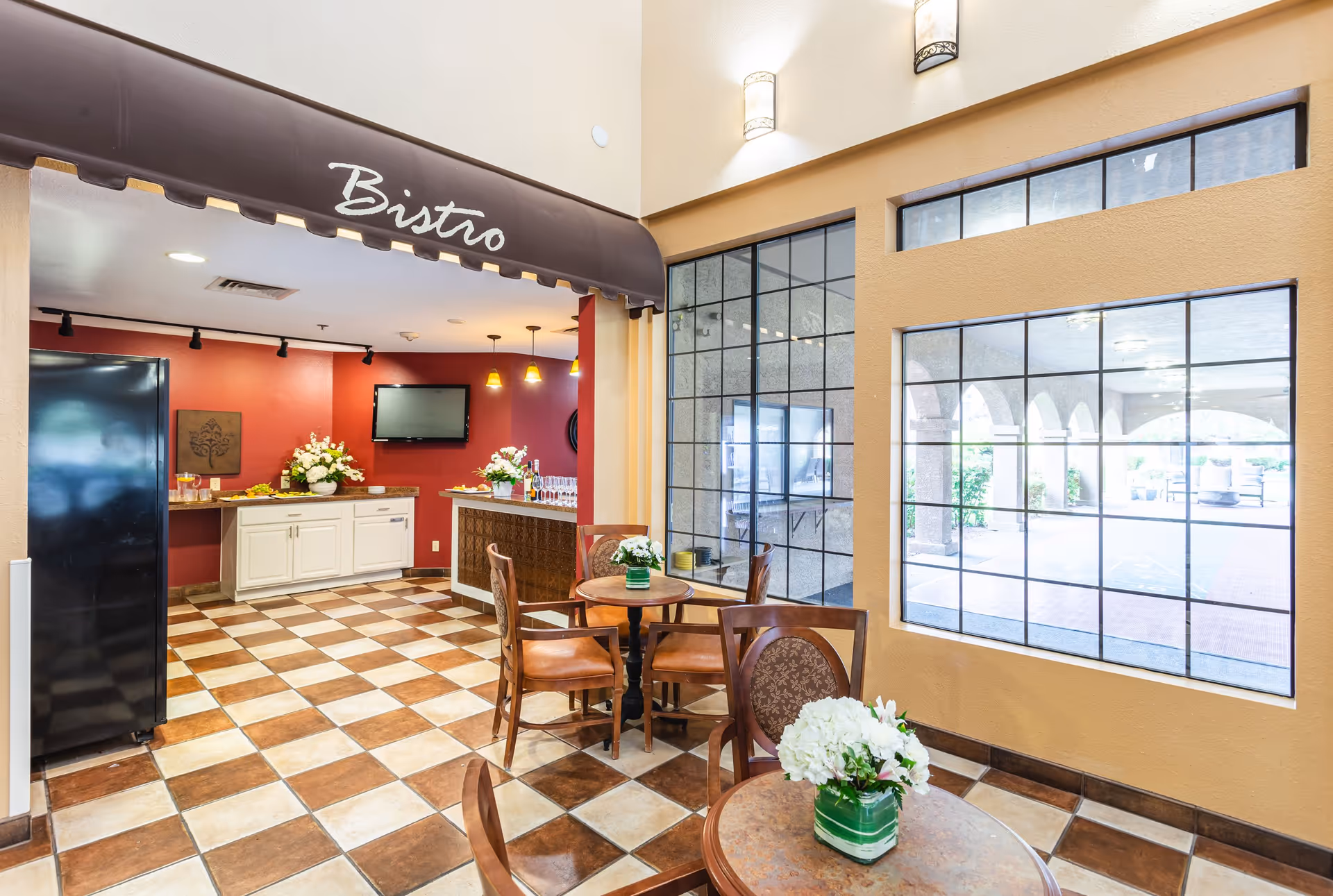 Interior view of a bistro area in a senior living facility named Garden Ridge, featuring a checkered tile floor, a small seating area with round tables and chairs, flower arrangements on the tables, a counter with white cabinets and a black refrigerator, and large windows letting in natural light.