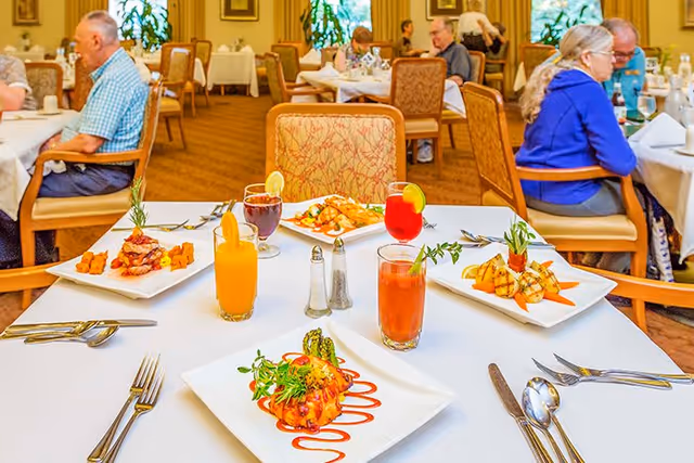 A dining room in a senior living facility with several tables set for meals. The foreground table has four plates of gourmet food and four colorful beverages. In the background, elderly people are seated at other tables, engaged in conversation and dining. The room is warmly lit with large windows and decorated with plants and framed pictures.