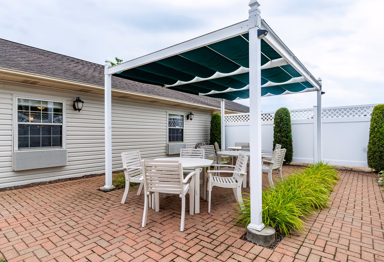 Outdoor patio area with white tables and chairs under a green fabric canopy. The patio is paved with red bricks and surrounded by a white fence and some green shrubs. The side of a beige building with windows and wall-mounted air conditioning units is visible.