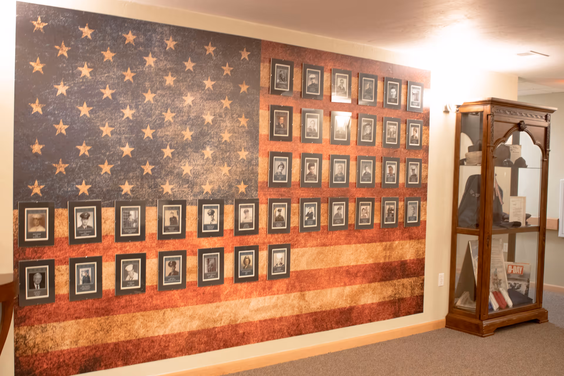 A wall decorated with a large American flag mural featuring framed black and white portraits of veterans. To the right, there is a wooden display cabinet containing various memorabilia and documents.