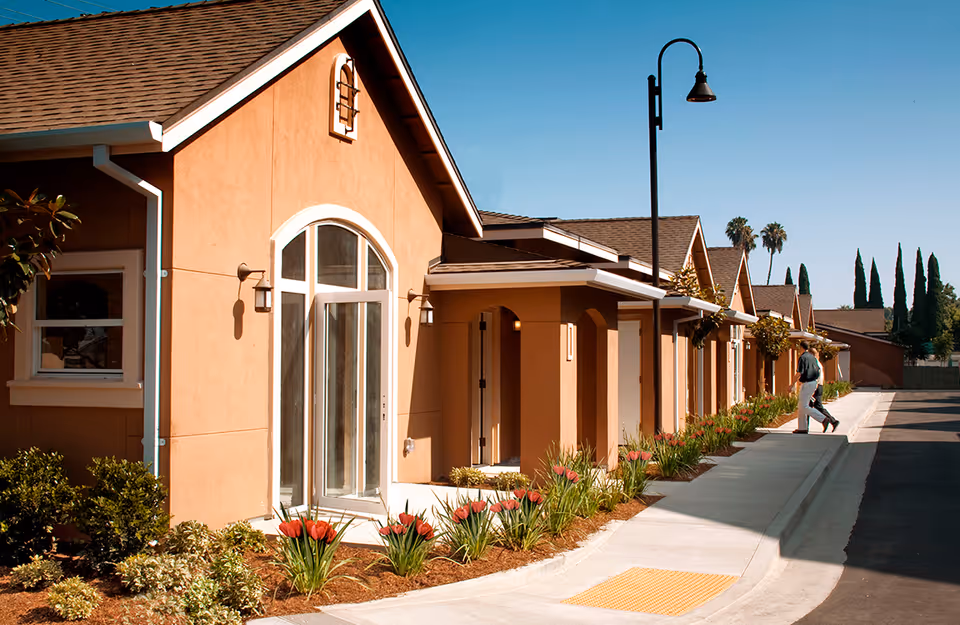 Row of single-story tan cottages at a retirement community with landscaped flower beds and a person walking on the sidewalk.