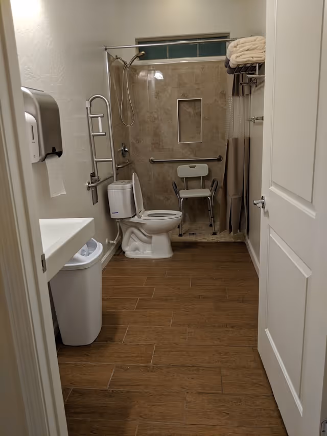 A bathroom with a toilet, a shower area with a shower chair, grab bars, a towel rack with folded towels, a sink with a paper towel dispenser above a trash bin, and a wooden floor.