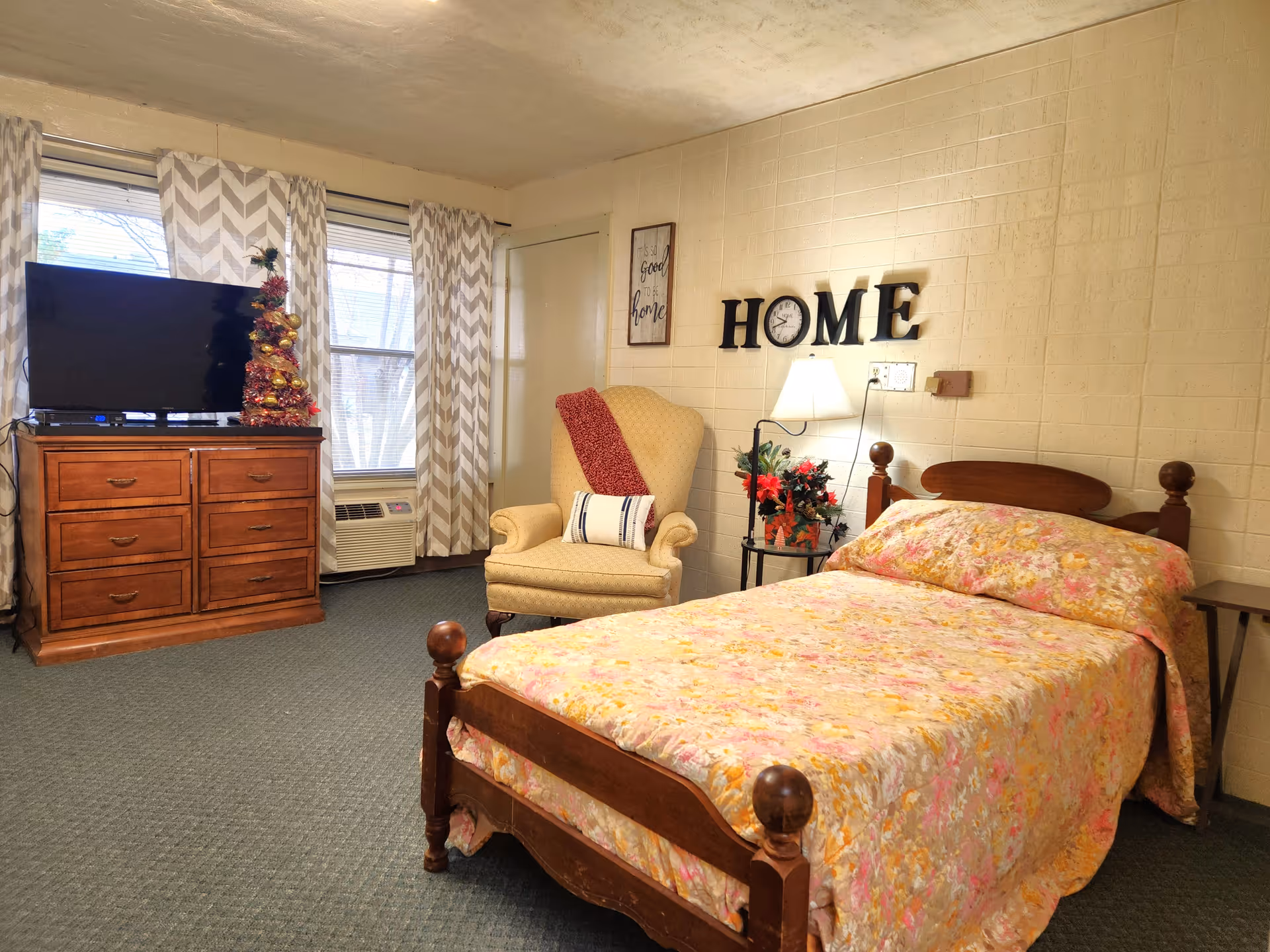 A cozy bedroom in an assisted living facility featuring a wooden bed with a floral bedspread, a cushioned armchair with a red throw, a wooden dresser with a flat-screen TV and a small decorated Christmas tree, two windows with chevron patterned curtains, and a wall decoration spelling 'HOME' with a lamp and a small table with flowers underneath.