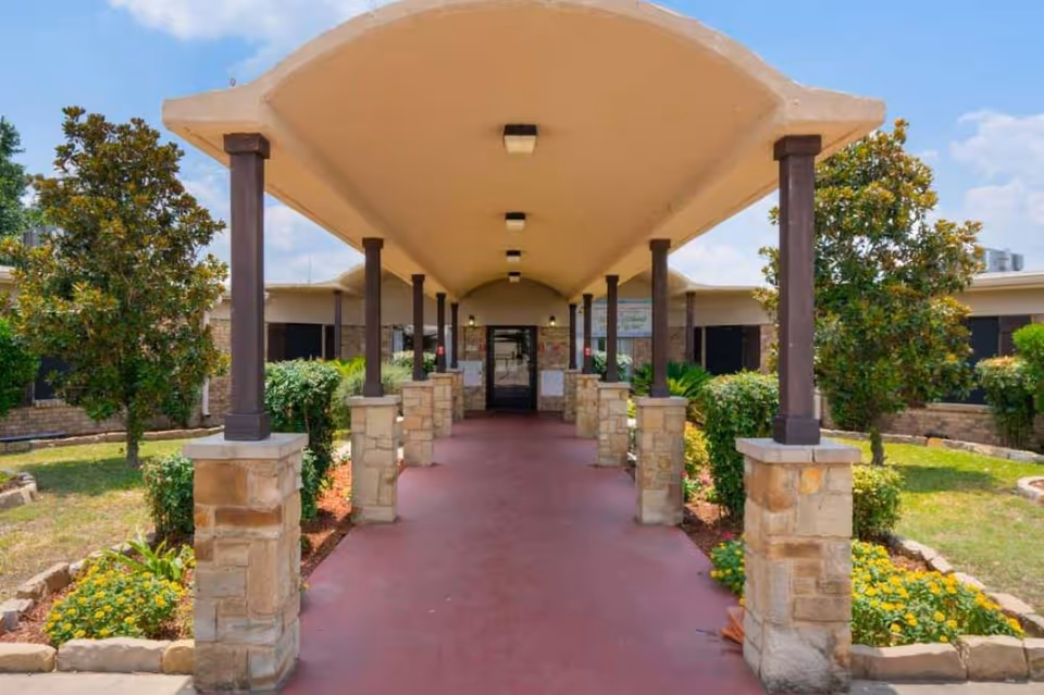 Covered walkway with stone pillars and brown columns leading to the entrance of a building surrounded by landscaped greenery and flowering plants under a partly cloudy sky.