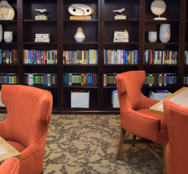 A cozy reading or common area with orange upholstered chairs around wooden tables, facing a large dark wooden bookshelf filled with books and decorative items such as vases and sculptures.