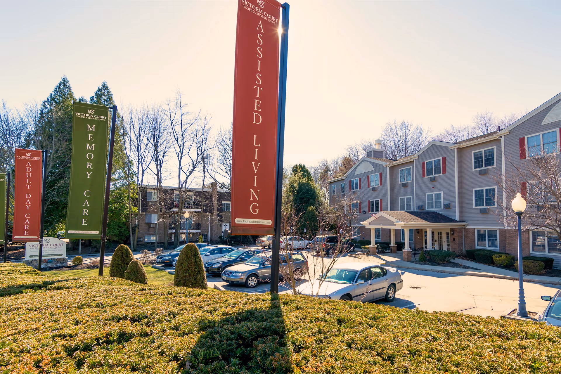 Front exterior of Victoria Court Memory Care assisted living facility showing large vertical signs, parked cars, and landscaped grounds.