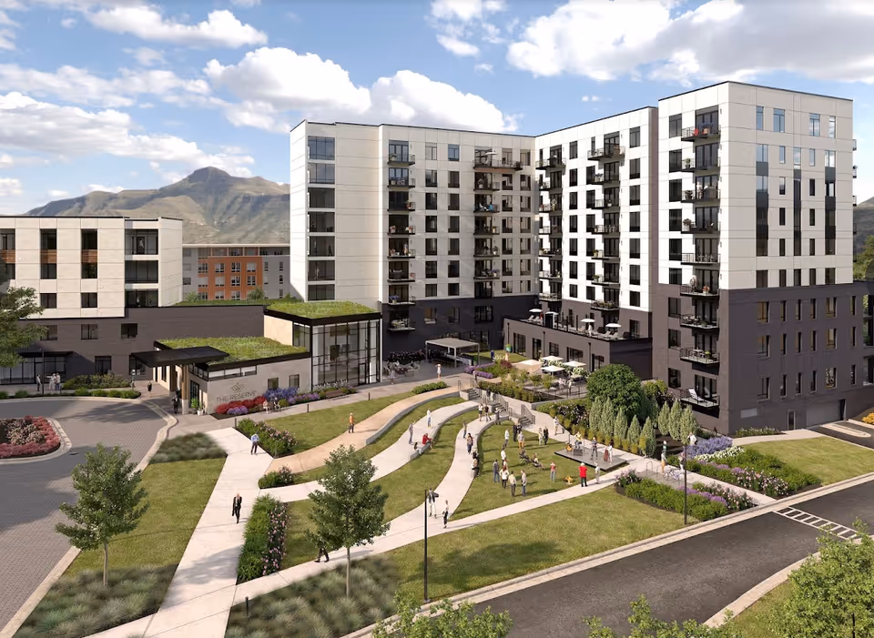 Exterior view of The Reserve at Lone Tree, a modern multi-story senior living facility with balconies. The building is surrounded by landscaped green lawns, walking paths, and outdoor seating areas. People are seen walking and socializing in the outdoor space. Mountains and a partly cloudy sky are visible in the background.