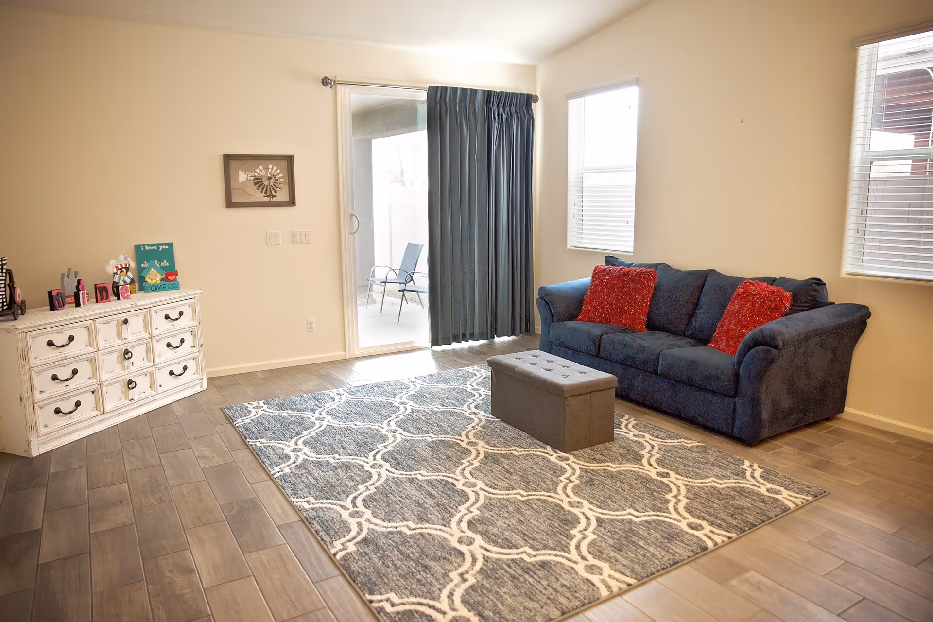 A bright living room with a dark blue couch adorned with two red pillows, a patterned area rug, a small gray ottoman, a white distressed dresser with decorative items, and sliding glass doors leading to an outdoor patio with chairs.