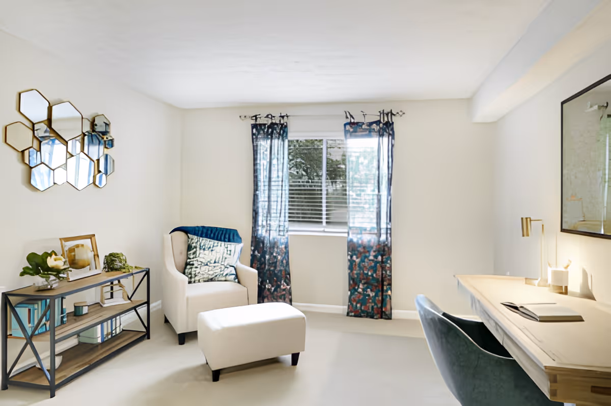 A bright and cozy living room with a beige armchair and matching ottoman near a window with blue patterned curtains. A wooden desk with a green chair and a lamp is on the right side. On the left, there is a metal and wood shelf with books and decorative items, and a hexagonal mirror arrangement on the wall above it.