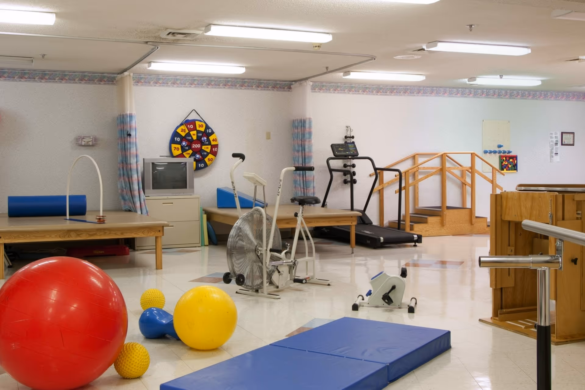 A rehabilitation center room with various physical therapy equipment including a large red exercise ball, yellow and blue therapy balls, an exercise bike, a treadmill, parallel bars, wooden stairs with handrails, a blue mat on the floor, and a dartboard on the wall above a TV and filing cabinet.