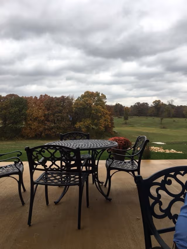 Wrought-iron patio table and chairs on a concrete patio overlooking a grassy field and trees under a cloudy sky.