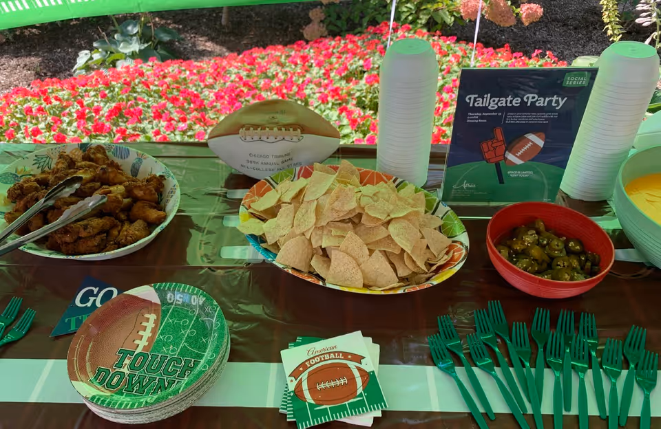 A table set up outdoors with a football-themed tailgate party spread including a bowl of chicken wings, a bowl of tortilla chips, a bowl of sliced jalapeños, a large bowl of cheese dip, green plastic forks, football-themed plates and napkins, and a sign advertising a Tailgate Party event. In the background, there are red flowers and greenery.