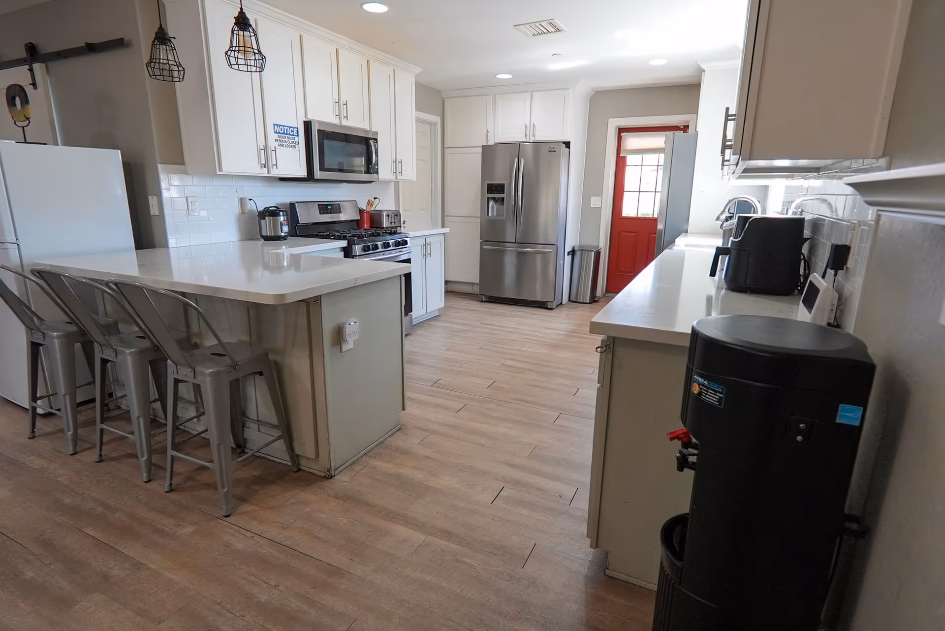 A modern kitchen with white cabinets, stainless steel refrigerator and stove, a microwave, and a white countertop island with four metal bar stools. The floor is wood-style tile, and there is a red door at the far end of the room. Pendant lights hang above the island, and various kitchen appliances are on the counters.