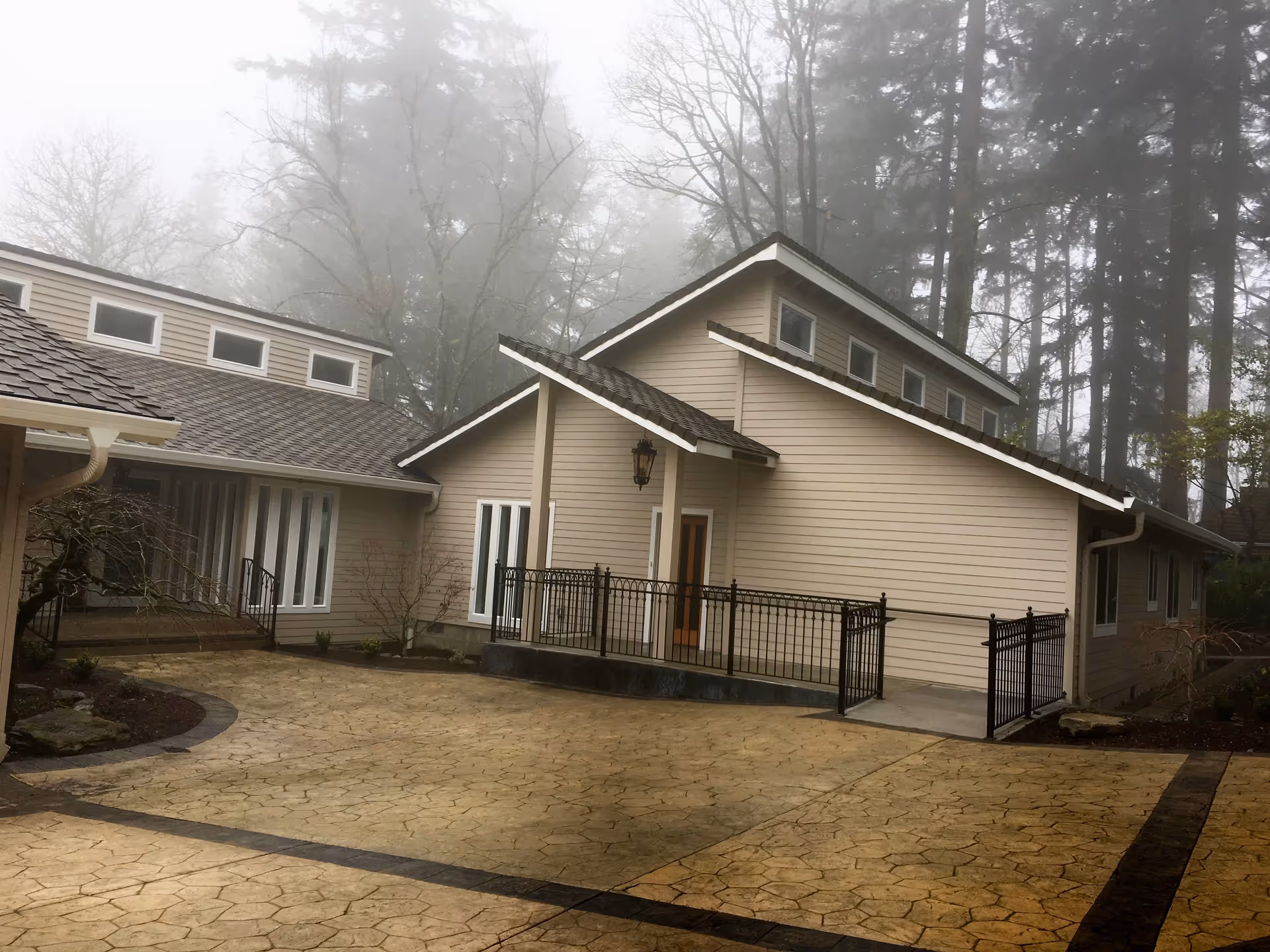 Exterior view of a beige single-story building with a paved courtyard, metal railing, and foggy trees in the background.